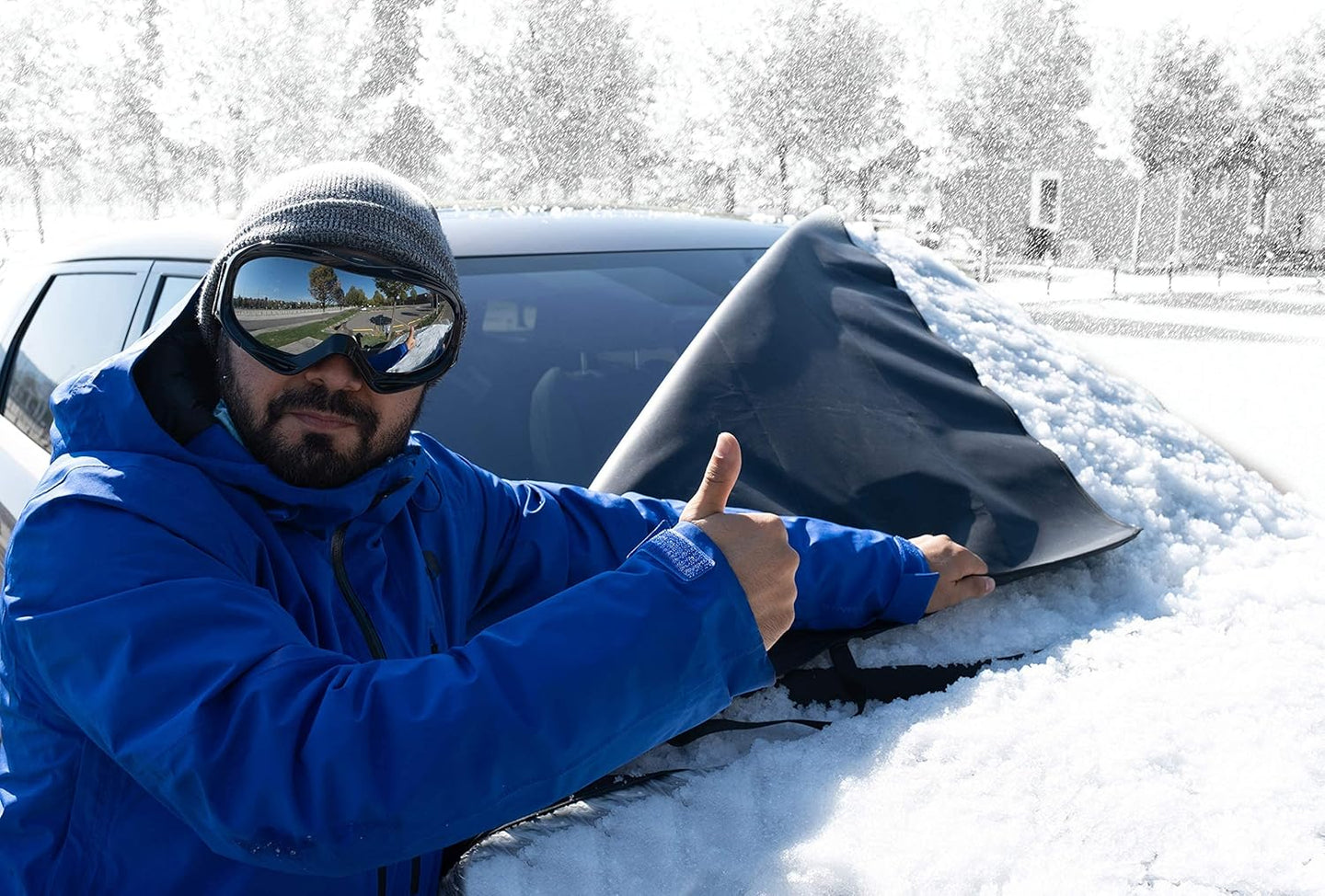 Person in blue jacket and goggles removing snow from a car window with a snow cover