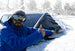 Person in blue jacket and goggles removing snow from a car window with a snow cover