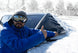 Person in blue jacket and goggles removing snow from a car window with a snow cover