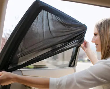 A woman installing a stretch-fit car window sunshade by sliding it over the side window frame to block sunlight.